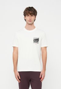 Biała bawełniana koszulka z grafiką czarno-białego oka na lewej kieszeni na piersi, w połączeniu z bordowymi spodniami.