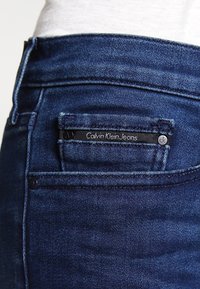 Jean en denim bleu foncé avec une poche arrière portant une étiquette noire inscrite "Calvin Klein Jeans" et un accent de bouton en métal.