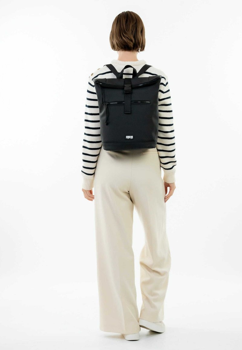 Sac à dos noir à rabat, avec des bretelles réglables, poche zippée à l'avant et une surface texturée. Le modèle porte un pull rayé et un pantalon crème.