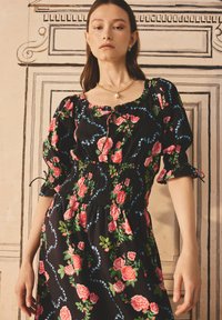 Rochie neagră cu flori, având trandafiri roz și frunze verzi, cu talie smocată și mâneci puff. Accentuată de un model spiralat albastru.