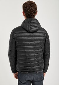 En person med lockigt hår bär en åtsittande svart pufferjacka med huva och står mot en enfärgad bakgrund, visande bakifrån.