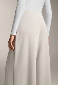 Wijdvallende beige broeken met een gladde textuur, met een hoge taille en subtiele stikdetails. Gecombineerd met een lichtgrijs shirt met lange mouwen.