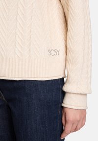 Krém színű, cable-knit stílusú pulóver bordás aljjal és mandzsettákkal, egy kisebb "SCsy" logós címkével. Sötét farmernadrággal párosítva.