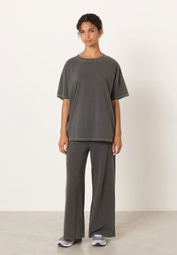 Mlada ženska z razmršenimi lasmi stoji, oblečena v ohlapno temno sivo majico in široke hlače, usklajene s sivimi supergami, na enobarvnem ozadju.