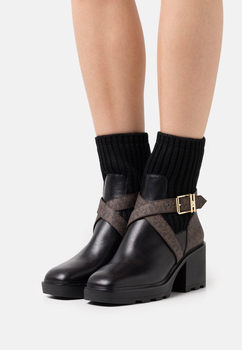 Bottines noires à talons blocs, brides croisées marron avec boucles dorées, portées avec des chaussettes en maille côtelée noire sur jambes nues.