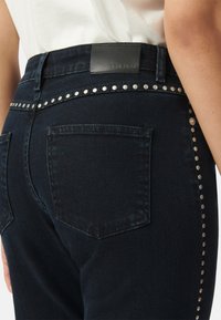 Jeans de mezclilla oscura con una etiqueta de cuero en la cinturilla, dos bolsillos traseros y una fila de tachuelas metálicas a lo largo de la cinturilla como acento.