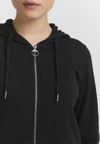 Felpa nera con zip realizzata in tessuto a costine. Caratterizzata da una zip frontale, tasca a marsupio e cordini con punte metalliche.