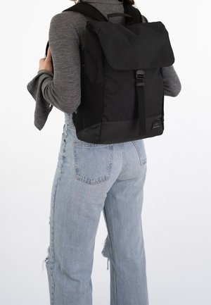 Svart ryggsäck i tyg med en övervikbar klaff, justerbara remmar och minimal hårdvara. Har en slät yta och kompakt design.