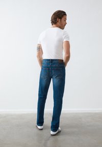 Jeans in denim blu con taglio dritto, tasche posteriori e una toppa in pelle marrone in vita, abbinati a una T-shirt bianca aderente.