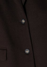 Brauner Wollmantel mit schmalem Revers, zwei Knöpfen und einer seitlichen Tasche aus glattem, strukturiertem Stoff.