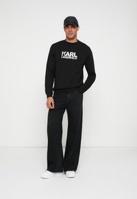 Svart sweatshirt med vitt "KARL LAGERFELD"-logo, kombinerad med svarta jeans med vida ben och vita sneakers; avslappnad look förstärkt med en svart keps.