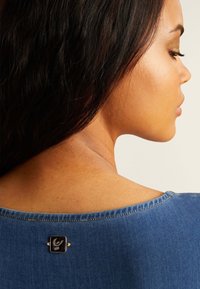 Top de mezclilla con un escote redondeado, que presenta un detalle de herrajes en negro y acentos dorados en la parte posterior, destacando la textura suave y las costuras limpias.