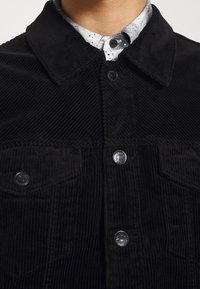 Chaqueta de pana negra con cierre de botones en la parte delantera, cuello puntiagudo y bolsillos delanteros con botones metálicos. La textura es acanalada y suave.