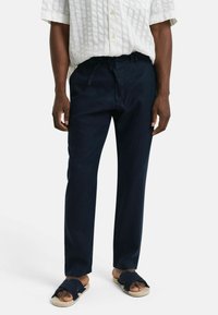 Navyblauwe linnen broek met een relaxte pasvorm, voorzien van een trekkoord in de taille, gecombineerd met zwarte sandalen met twee banden op een lichte achtergrond.