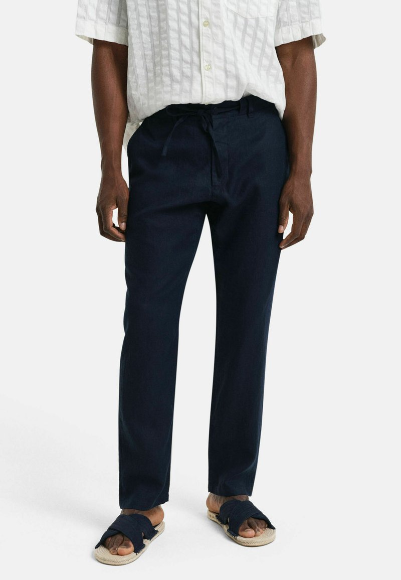 Navyblauwe linnen broek met een relaxte pasvorm, voorzien van een trekkoord in de taille, gecombineerd met zwarte sandalen met twee banden op een lichte achtergrond.