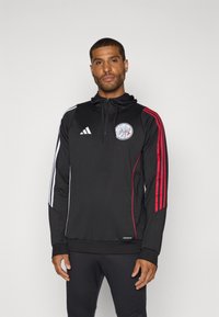 Schwarze sportliche Kapuzenjacke aus glattem Stoff. Mit roten und weißen Streifen an den Ärmeln, einem Logo auf der Brust sowie geripptem Saum und Bündchen.