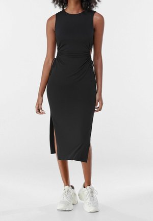 Femme vêtue d'une robe midi noire sans manches avec une fente latérale et des baskets blanches épaisses, debout devant un fond blanc uni.