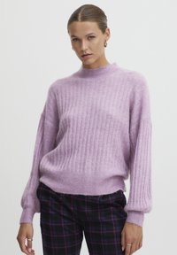 Maglione lavanda a coste con collo alto e maniche voluminose, realizzato in un materiale morbido e testurizzato. Abbinato a pantaloni a quadri.