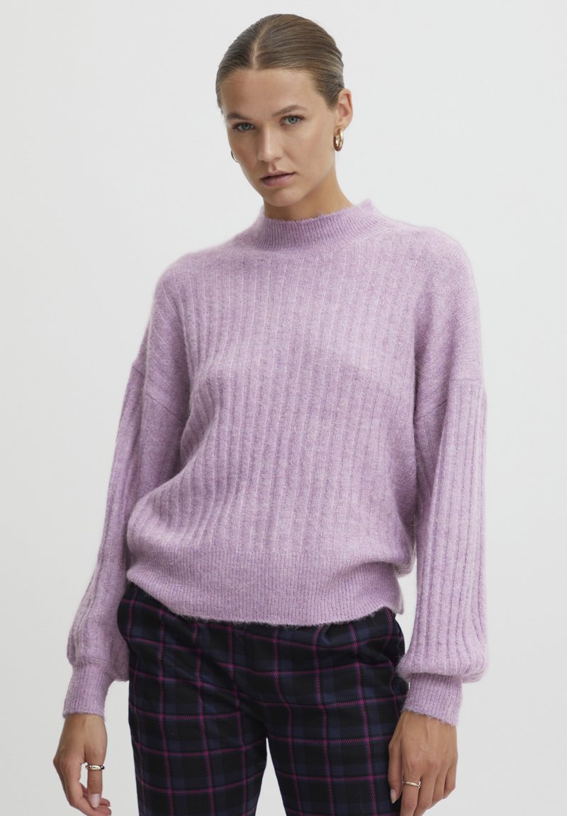Maglione lavanda a coste con collo alto e maniche voluminose, realizzato in un materiale morbido e testurizzato. Abbinato a pantaloni a quadri.