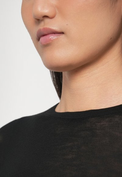 Top nero in maglia con scollo rotondo, caratterizzato da una texture morbida e dettagli di cuciture visibili sul colletto. Pelle liscia tono neutro sotto il colletto.