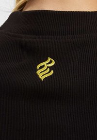 Svart ribbet stoff med et markant gult brodert logo, plassert på baksiden nær halslinningen.