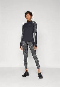 Top negro de manga larga con media cremallera y detalles reflectantes, combinado con leggings ajustados en negro y gris con estampado. Zapatillas deportivas negras.