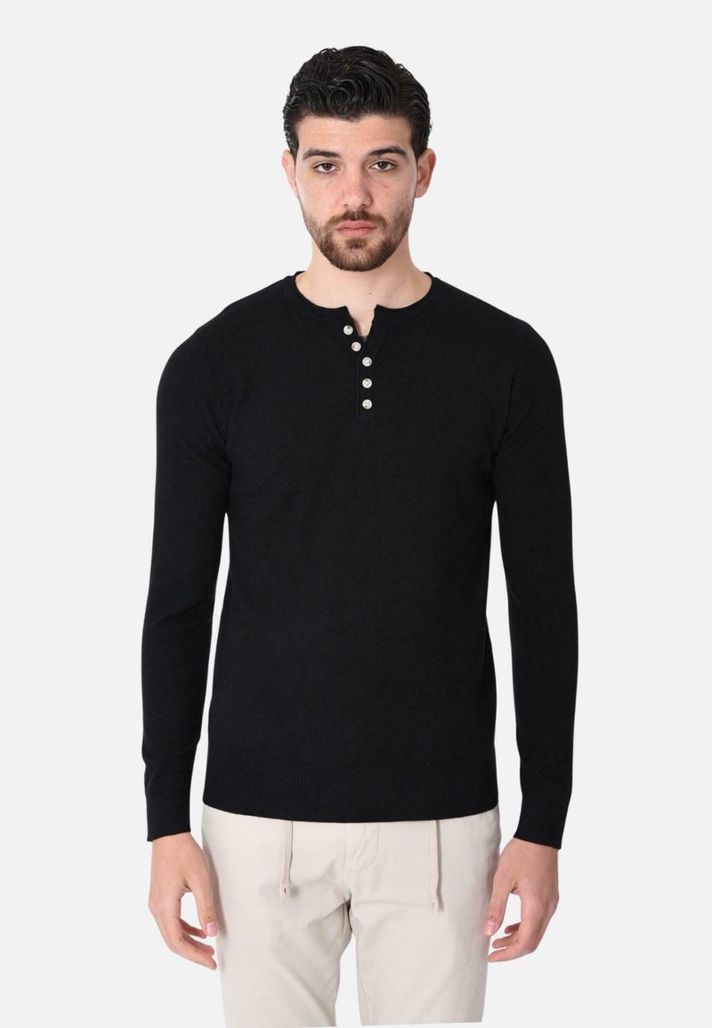 Maglione henley a maniche lunghe in nero con un motivo testurizzato, patta con cinque bottoni e polsini a coste. Abbinato a pantaloni chiari.