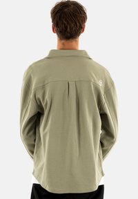 Camisa de manga larga en color verde oliva con un tejido texturizado, cuello tipo polo, detalle de pliegue en la parte trasera y logo bordado en la parte superior derecha.
