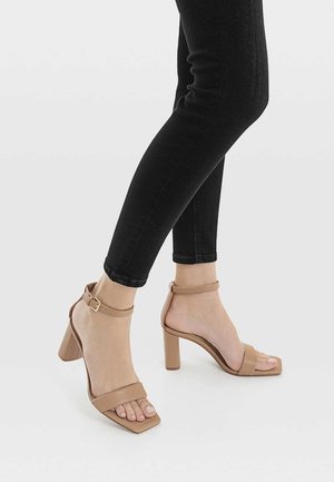 Sandales à talons beiges avec un design à bout carré. Dispose de deux larges lanières sur le pied et d'une lanière de cheville avec boucle. Texture lisse.