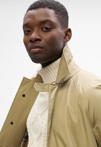 Beige, op maat gemaakte trenchcoat met een hoge kraag, voorzien van een soepele textuur en subtiele glans. Gelaagd over een crème kabelgebreide trui.