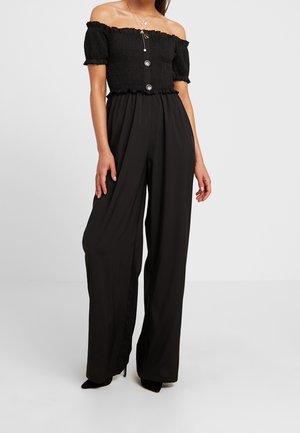 Femme portant un haut noir smocké à épaules dénudées avec des boutons, un pantalon noir taille haute à jambes larges et des talons noirs.