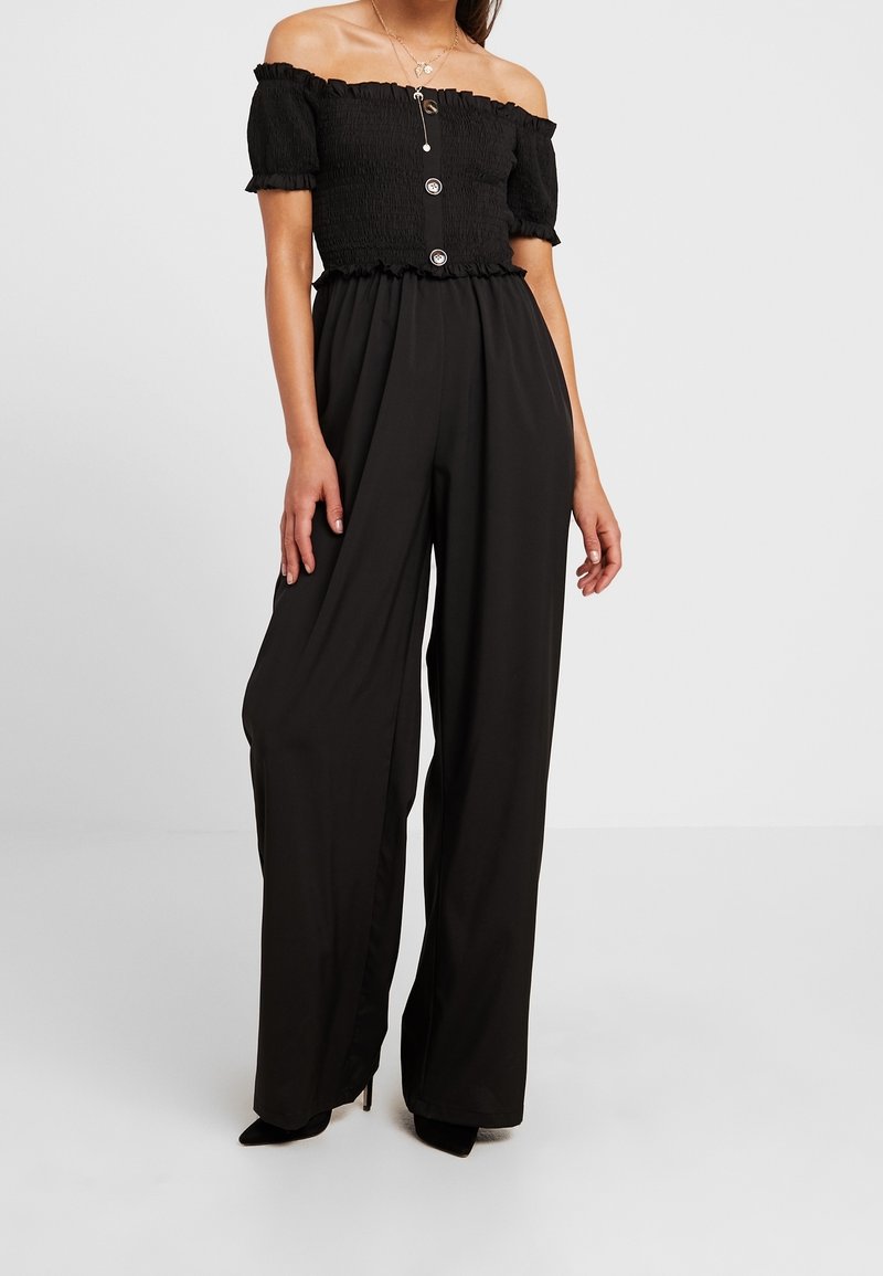 Femme portant un haut noir smocké à épaules dénudées avec des boutons, un pantalon noir taille haute à jambes larges et des talons noirs.