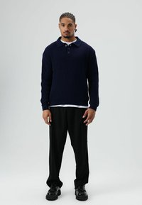 Jonge man met gevlochten haar, draagt een marineblauwe gebreide trui, zwarte wijde broek en zwarte schoenen, staand tegen een effen grijze achtergrond.