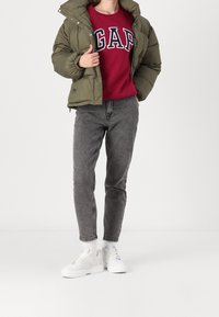 Prošívaná bunda olivově zelené barvy s vysokým límcem, nošená přes bordó mikinu GAP. Šedé džínové kalhoty a bílé tenisky na platformě dokončují vzhled.