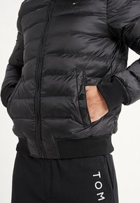 Veste matelassée noire avec fermeture éclair, motif matelassé horizontal, col montant et poignets côtelés. Comprend des poches latérales avec bouton-pression.