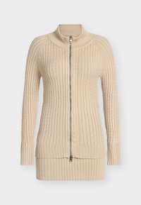 Beige stickad tröja med hög krage och hel dragkedja, med kabelstickade mönster samt ribbstickade muddar och fåll. Mjuk textur.