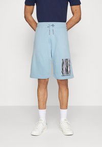 Lichtblauwe shorts van zachte stof, tot op de knie, met een elastische tailleband en trekkoord. Heeft een zwart-wit grafisch ontwerp op de linkerdij.