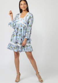 Mujer de pie con un vestido floral azul con encaje y mangas largas, usando sandalias de tacón beige contra un fondo liso.