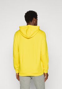 Gul hættetrøje med en blød tekstur, der har en rummelig hætte og ribbede manchetter, båret over grå sweats.