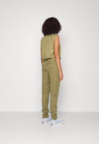Top sin mangas de color verde oliva combinado con pantalones taperados a juego, que presentan una cintura elástica y bolsillos tipo deslizamiento, llevados con zapatillas de color azul claro.