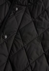 Sort quiltet jakke med diamantmønster, der har en rund sort knap og en broderet logo detajle. Glat tekstur.
