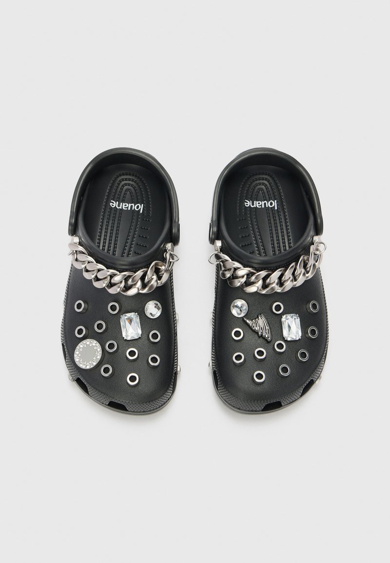 Schwarze Clogs mit strukturiertem Design, metallischen Kettenakzenten, dekorativen Edelsteinen und runden Ausschnitten im Obermaterial.