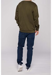 Olivgrön bomulls-tröja med rund halsringning och ribbade ärmslut, matchad med mörkblå jeans och vita sneakers med röda och marinblå accenter.