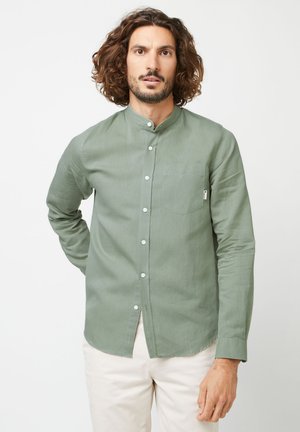 Mann mit mittellangem lockigem Haar, der ein hellgrünes Hemd mit Stehkragen und beige Hose trägt, vor einem weißen Hintergrund stehend.