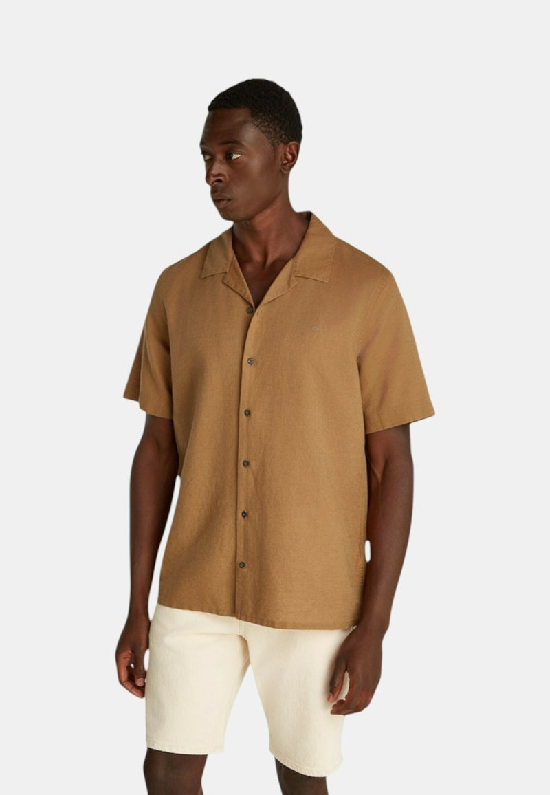 Camicia button-up in lino marrone a maniche corte con colletto piatto, caratterizzata da un logo sul petto discreto e cinque bottoni scuri. Abbinata a pantaloni corti color crema.
