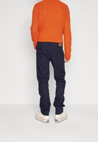 Maglione arancione lavorato a maglia, jeans blu scuro e sneakers bianche con accenti neri; anello per cintura visibile e due tasche posteriori sui jeans.