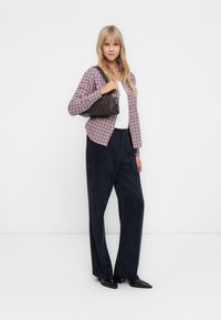 Chemise à carreaux dans des tons violets, haut blanc, pantalon large foncé et sac à main sombre. Le modèle se tient sur un fond uni.
