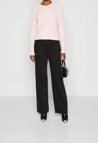 Vaaleanpunainen teksturoidusta materiaalista valmistettu neule, jossa on pitkät hihat, yhdistetty korkeavyötäröisten mustien leveälahkeisten housujen ja mustien kärjellisten korkokenkien kanssa. Pieni käsilaukku.