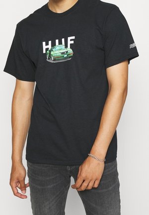 Osoba ubrana w czarną koszulkę z napisem „HUF” oraz zielonym nadrukiem samochodu na przodzie, w zestawieniu z ciemnoszarymi dżinsami i srebrną bransoletką.