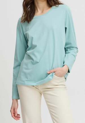 Femme portant une chemise à manches longues bleu clair et un pantalon crème, avec une main dans la poche, sur un fond uni.